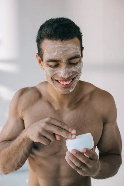 Musculoso hombre de raza mixta con crema en la cara tomando crema cosmética con los dedos del envase - foto de stock