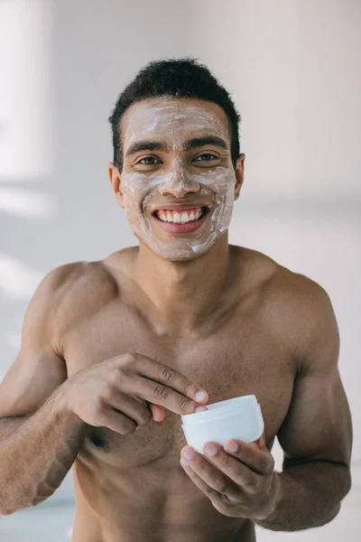 Muscoloso uomo di razza mista con crema sul viso che prende crema cosmetica con le dita dal contenitore mentre guarda la fotocamera — Foto stock