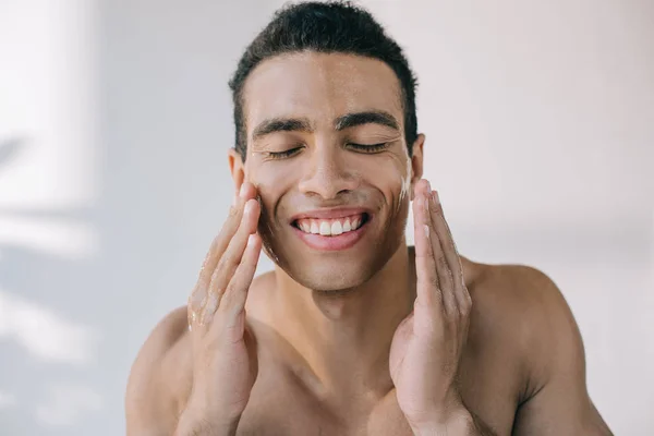 Beau jeune homme au visage mouillé le touchant avec les mains tout en souriant avec les yeux fermés — Photo de stock