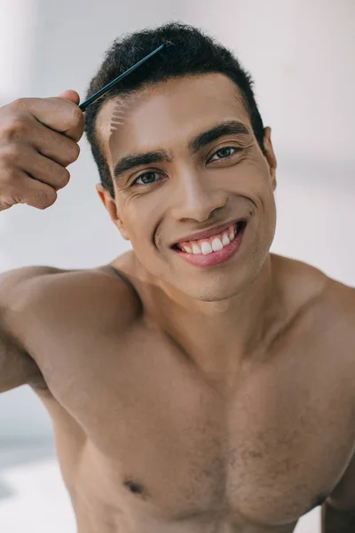 Guapo hombre de raza mixta peinando el cabello con cepillo y sonriendo mientras mira la cámara - foto de stock
