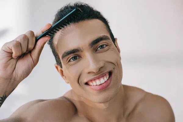 Retrato de hombre de raza mixta peinando el cabello con cepillo y sonriendo mientras mira a la cámara - foto de stock