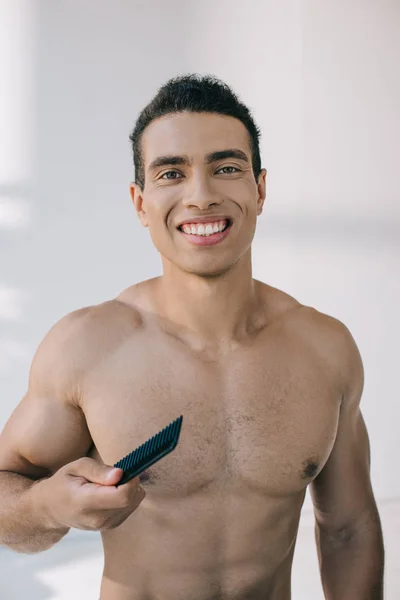 Guapo mixto raza hombre sosteniendo cepillo de pelo y sonriendo mientras mira a la cámara - foto de stock