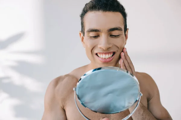 Handsome mixed race man touching face with hand while smiling and looking at mirror — Stock Photo