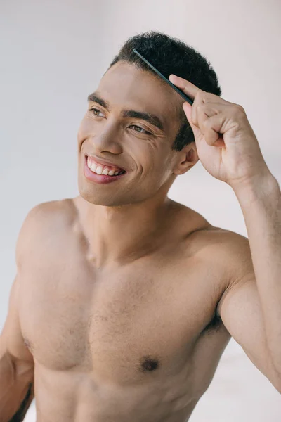 Bonito misto raça homem pentear o cabelo com escova de cabelo e sorrindo enquanto olha para longe — Fotografia de Stock