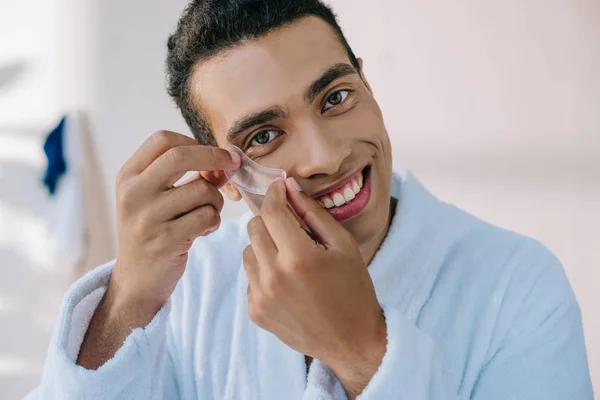 Handsome young man in bathrobe putting patch under eye and smiling while looking at camera — Stock Photo