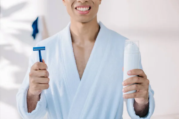 Vista recortada del hombre en albornoz celebración de la maquinilla de afeitar y dispensador con crema de afeitar - foto de stock