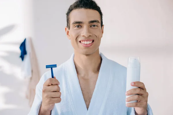 Joven guapo en albornoz sosteniendo maquinilla de afeitar y dispensador con crema de afeitar mientras mira a la cámara - foto de stock