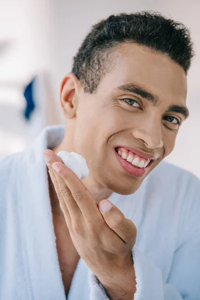 Retrato de hombre guapo aplicando crema de afeitar en la cara mientras mira a la cámara - foto de stock