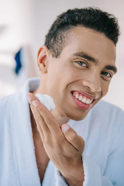 Retrato tiro de homem bonito aplicando creme de barbear no rosto enquanto olha para a câmera — Fotografia de Stock