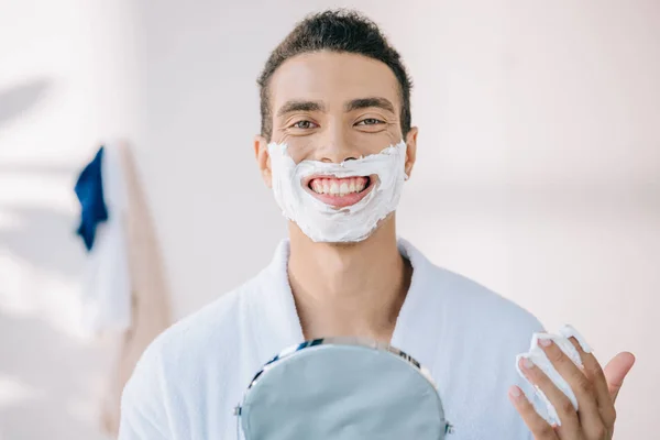 Handsome young man in bathrobe with shaving cream on face holding mirror while smiling and looking at camera — Stock Photo