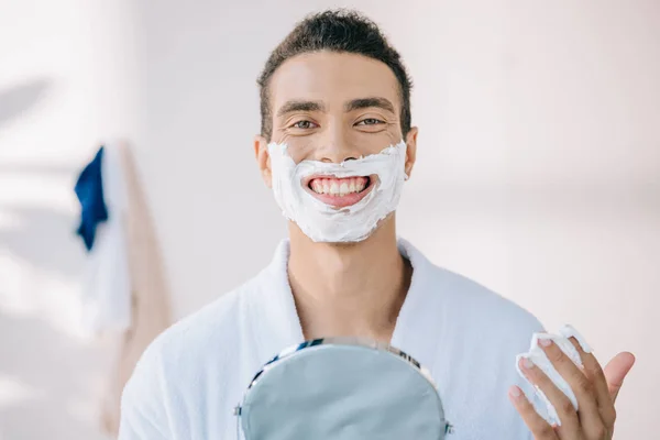 Beau jeune homme en peignoir avec crème à raser sur le visage tenant miroir tout en souriant et en regardant la caméra — Photo de stock