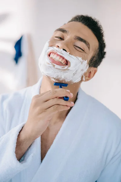 Retrato de un hombre guapo afeitándose la cara con una navaja mientras sonríe y mira a la cámara - foto de stock