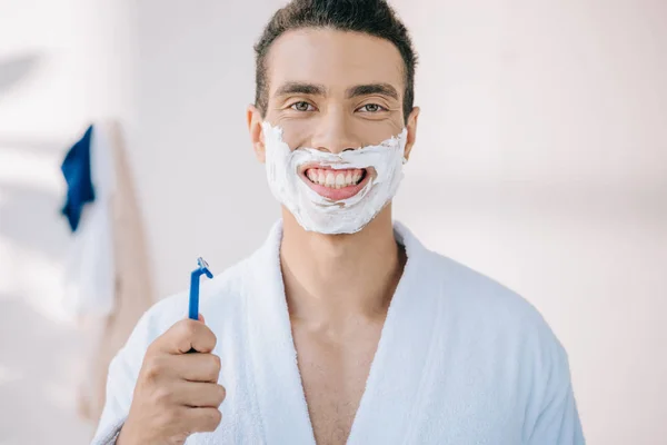 Красивый мужчина с кремом для бритья на лице, держащий бритву и смотрящий в камеру — стоковое фото