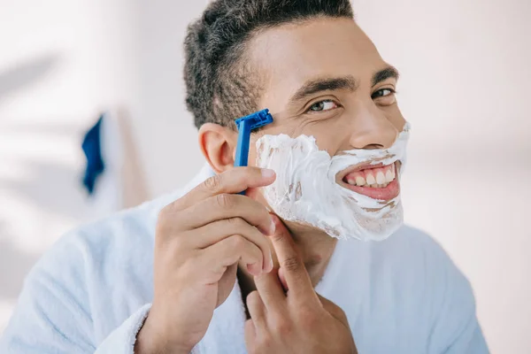 Portrait shot of handsome man in bathrobe shaving face with razor while smiling and looking at camera — Stock Photo