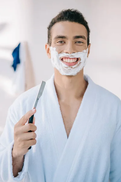 Jovem bonito com creme de barbear na cara segurando lâmina de barbear, sorrindo e olhando para a câmera — Fotografia de Stock