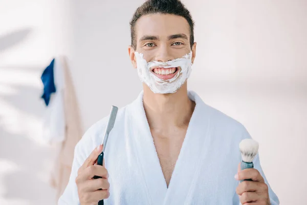 Joven guapo con crema de afeitar en la cara sosteniendo hoja de afeitar y cepillo de afeitar, sonriendo y mirando a la cámara - foto de stock
