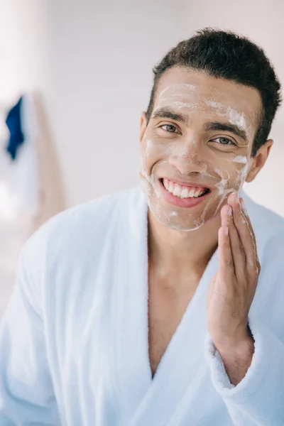 Schöner junger Mann im Bademantel, der Schaum aufs Gesicht sprüht und in die Kamera lächelt — Stockfoto