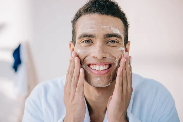 Handsome young man in bathrobe applying facial foam with hands and smiling while looking at camera — Stock Photo