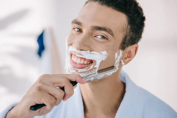 Handsome young man in bathrobe shaving face with razor blade, smiling and looking at camera — Stock Photo