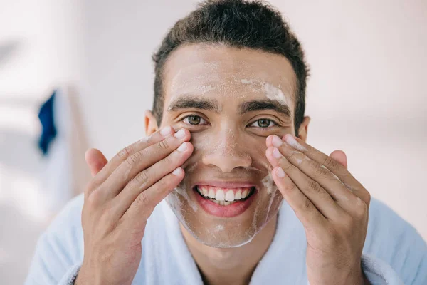 Handsome young man in bathrobe washing face with facial foam and smiling while looking at camera — Stock Photo