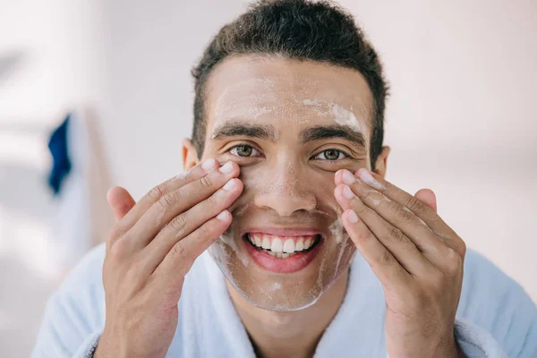 Joven guapo en la cara de lavado de albornoz con espuma facial y sonriendo mientras mira a la cámara - foto de stock