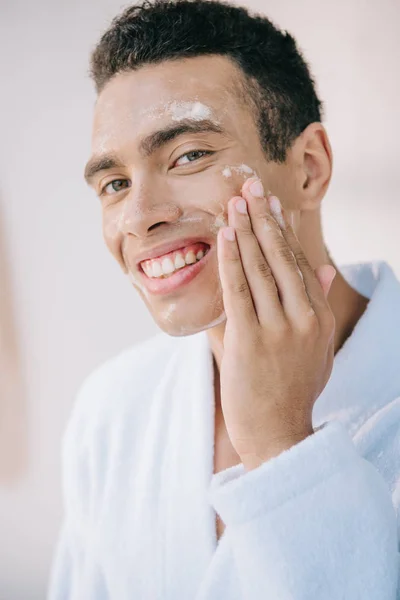 Schöner junger Mann im Bademantel wäscht Gesicht mit Schaum und lächelt in die Kamera — Stockfoto