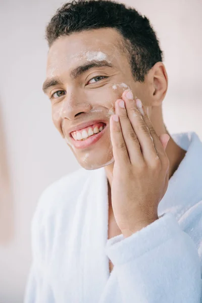 Joven guapo en albornoz lavando la cara con espuma y sonriendo mientras mira a la cámara - foto de stock