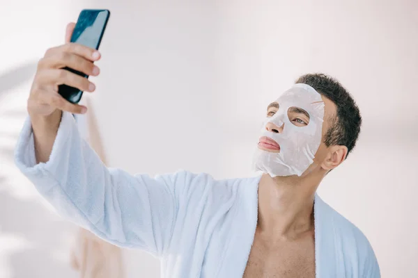 Jeune homme en peignoir avec masque cosmétique sur le visage prenant selfie sur smartphone — Photo de stock