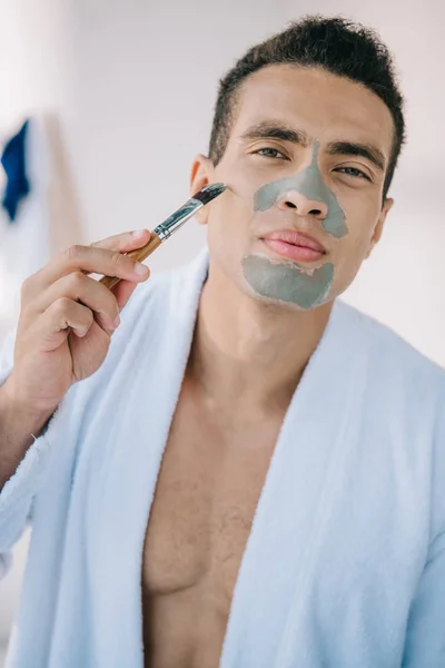 Schöner Mann, der im Bademantel mit Pinsel Tonmaske auf Gesicht aufträgt — Stockfoto