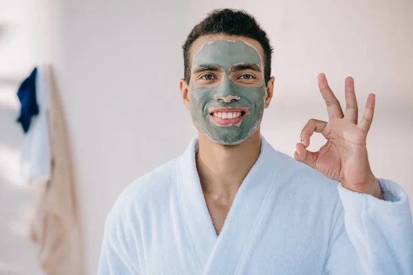 Schöner Mann mit Gesichtsmaske und im Bademantel mit Okay-Zeichen und Blick in die Kamera — Stockfoto