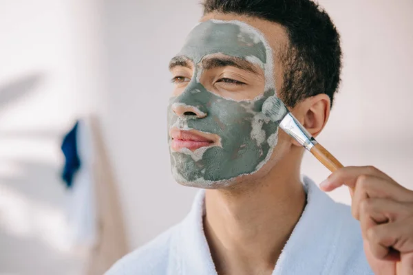 Bonito homem aplicando cinza máscara facial com escova e olhando para longe — Fotografia de Stock