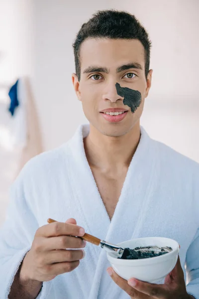 Schöner junger Mann im Bademantel hält Schale mit Gesichtsmaske und trägt sie mit Pinsel auf das Gesicht auf — Stockfoto