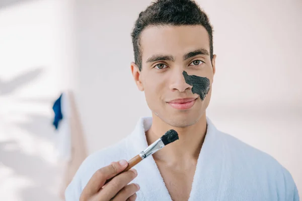 Beau jeune homme en peignoir tenant bol avec masque facial et l'appliquant sur le visage avec une brosse tout en regardant la caméra — Photo de stock