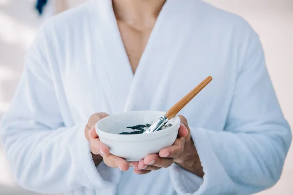 Cropped view of handsome young man in bathrobe holding bowl with face mask and brush — Stock Photo