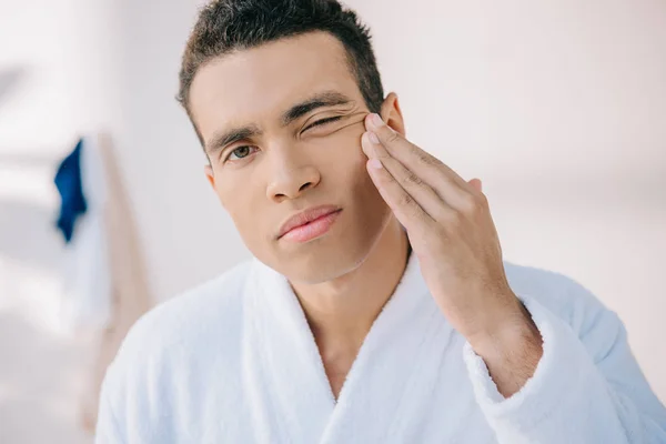 Sérieux jeune homme en peignoir toucher le visage et regarder la caméra — Photo de stock