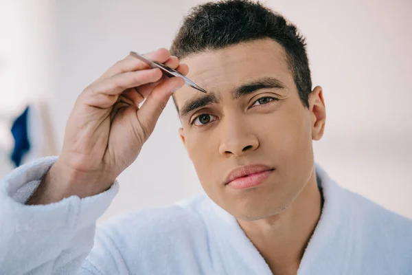 Portrait shot of handsome man plucking eyebrows with tweezer and looking at camera — Stock Photo