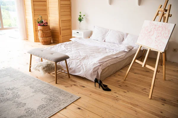 Спальня з білою ковдрою та подушками, мольберт, приліжкова лавка, килим, пристрій кімнати та чорні підбори на дерев'яній підлозі — стокове фото