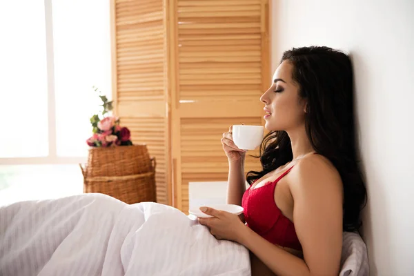 Vista lateral de hermosa chica sexy sentada en la cama con los ojos cerrados y beber café - foto de stock