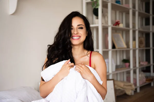 Сексуальная девушка в красном нижнем белье сидит в постели, покрытая одеялом, улыбается и смотрит в камеру — стоковое фото