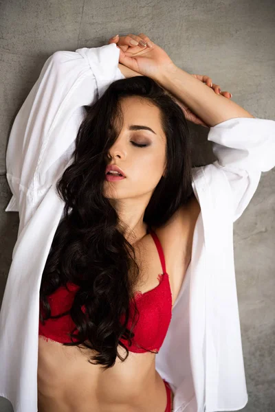 Retrato de chica sexy en ropa interior roja y camisa blanca apoyada en la pared gris con la mano sobre la cabeza y con los ojos cerrados - foto de stock