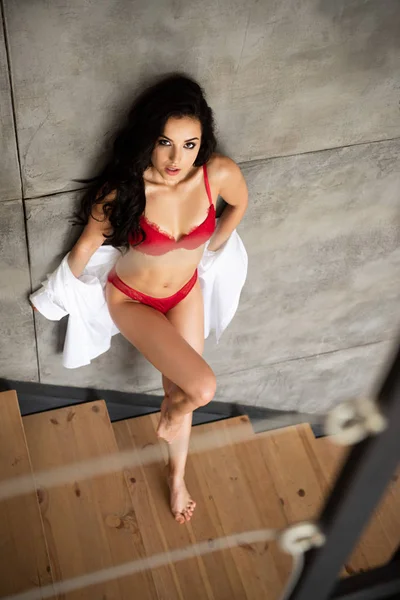Vista ad alto angolo della ragazza sexy in biancheria intima rossa e camicia bianca appoggiata sulla parete grigia e guardando la fotocamera — Foto stock