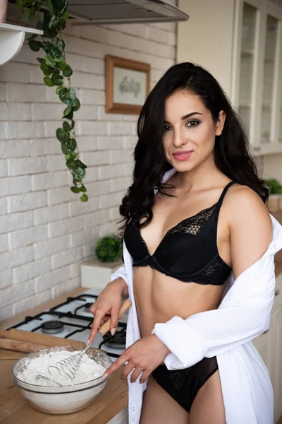 Menina sexy em roupa interior preta e camisa branca fazendo massa enquanto estava na cozinha e olhando para a câmera — Fotografia de Stock