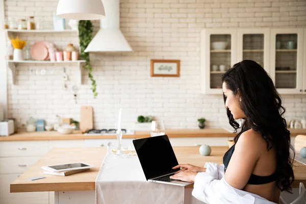 Vista trasera de chica sexy en ropa interior negra y camisa blanca usando el ordenador portátil mientras está sentado en la cocina - foto de stock