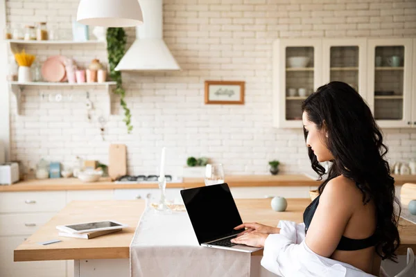 Back view of sexy girl in black underwear and white shirt using laptop while sitting in kitchen — Stock Photo