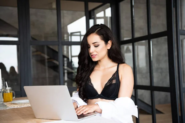 Sexy girl in black underwear and white shirt using laptop while sitting in kitchen — Stock Photo