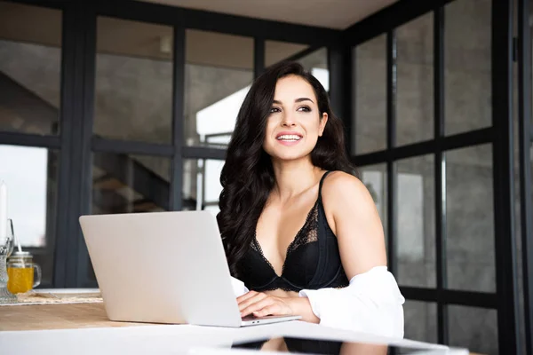 Chica sexy en ropa interior negra y camisa blanca usando el ordenador portátil mientras está sentado en la cocina y mirando hacia otro lado - foto de stock