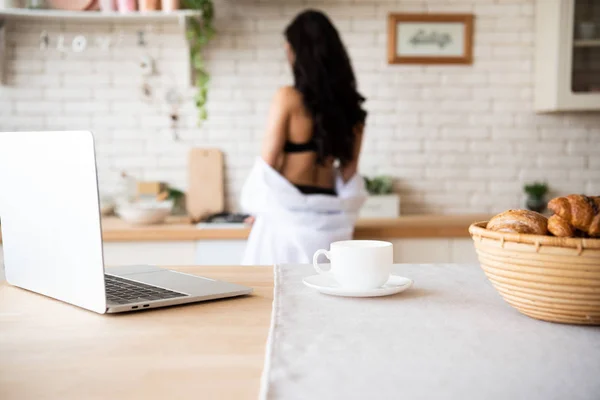 Селективный фокус ноутбука, чашки кофе и корзины с булочками на столе с девушкой на заднем плане — стоковое фото