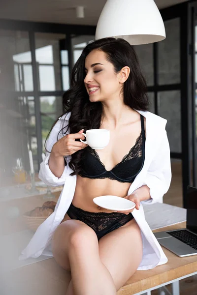Сексуальная девушка в черном нижнем белье и белой рубашке пьет кофе с закрытыми глазами, сидя на столе на кухне — стоковое фото