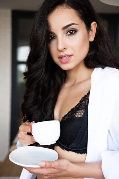 Портрет сексуальной девушки в черном белье и белой рубашке, пьющей кофе и смотрящей в камеру — стоковое фото