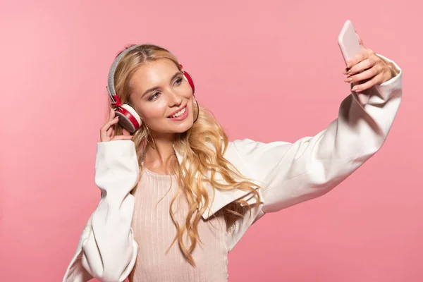 Beautiful blonde woman with headphones taking selfie on smartphone isolated on pink — Stock Photo